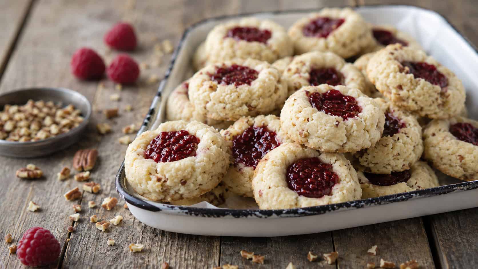 pale pecan sandies with raspberry-filled centers on a rustic enamel tray, in a wide horizontal shot, chopped pecans and a few raspberries scattered on a weathered wood table, soft natural light, shallow depth of field, landscape orientation