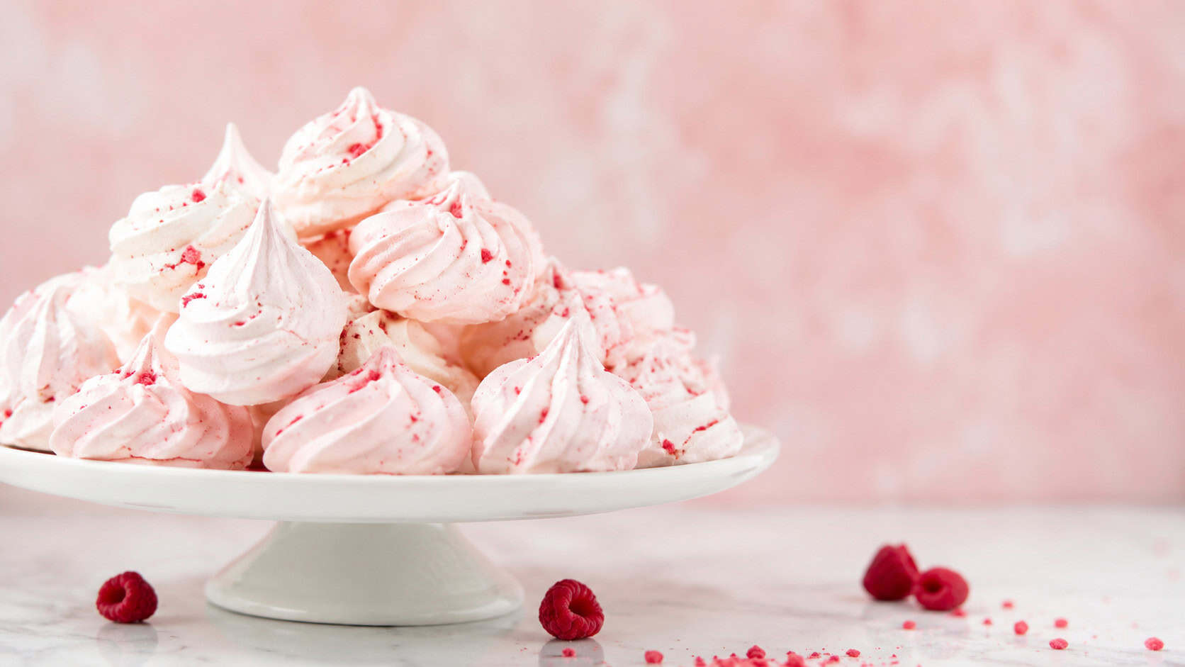 delicate pale pink meringue clouds piled on a white cake stand, in a wide horizontal shot, a few crushed raspberries and a soft pink backdrop, bright airy natural light, shallow depth of field, landscape orientation