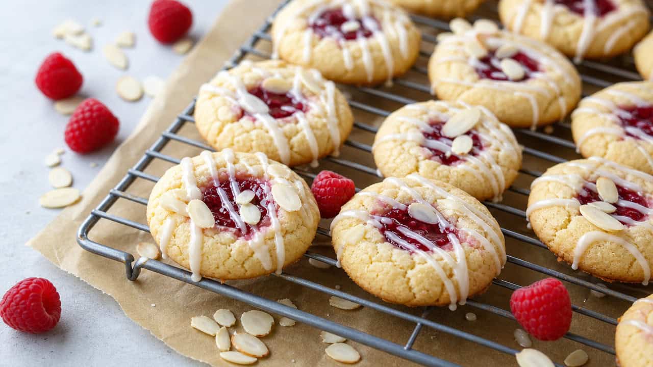 soft almond cookies with raspberry centers and white glaze drizzle on a cooling rack over parchment, in a wide horizontal shot, a few whole raspberries and sliced almonds scattered around, bright natural light, shallow depth of field, landscape orientation