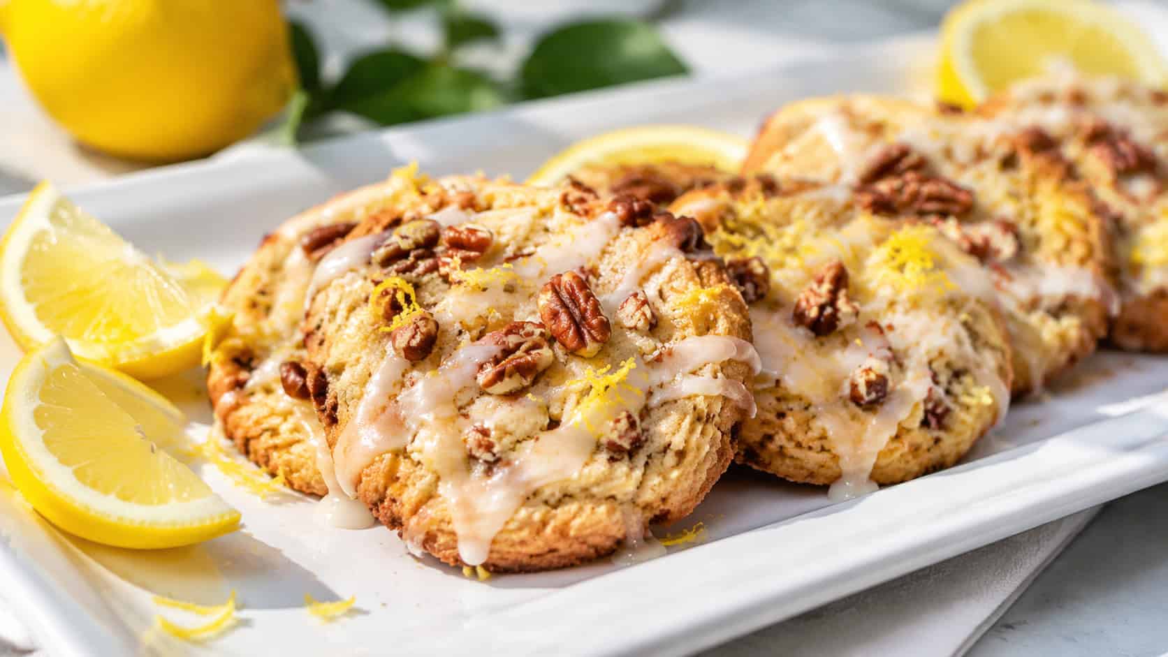 glazed lemon pecan cookies in a wide horizontal shot, drizzled with white glaze and garnished with lemon zest, arranged on a bright white platter with lemon wedges nearby, sunny natural light, shallow depth of field, landscape orientation