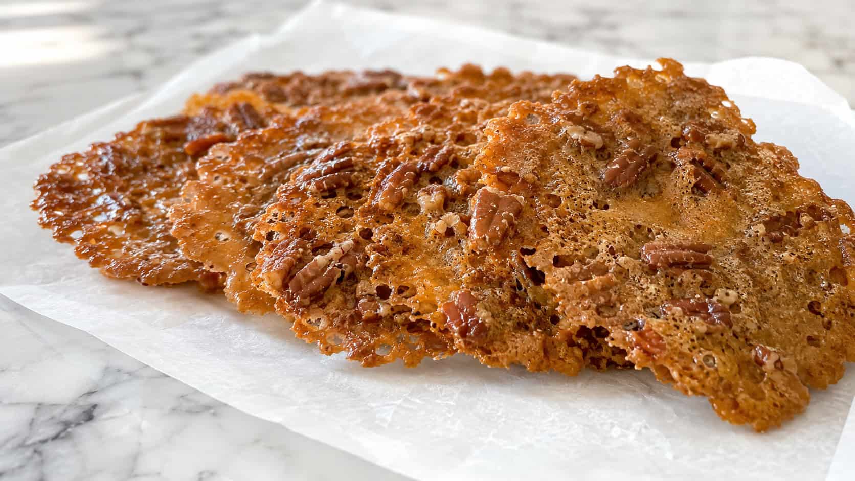 thin lacy pecan praline crisps in a wide horizontal shot, caramel-golden and translucent at the edges, fanned out on a sheet of parchment over a marble counter, bright natural light, shallow depth of field, landscape orientation