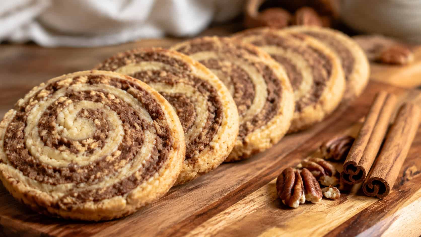 pinwheel cinnamon pecan swirl cookies in a wide horizontal shot, showing spiral patterns, lined up on a wooden cutting board with cinnamon sticks beside them, warm natural light, shallow depth of field, landscape orientation