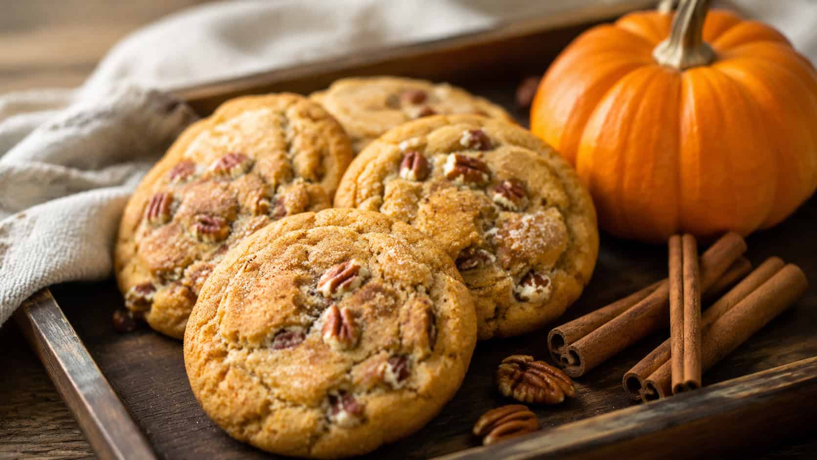 soft pumpkin pecan spice cookies in a wide horizontal shot, golden-orange and pillowy, arranged on a rustic tray with a small pumpkin and cinnamon sticks alongside, warm natural light, shallow depth of field, landscape orientation