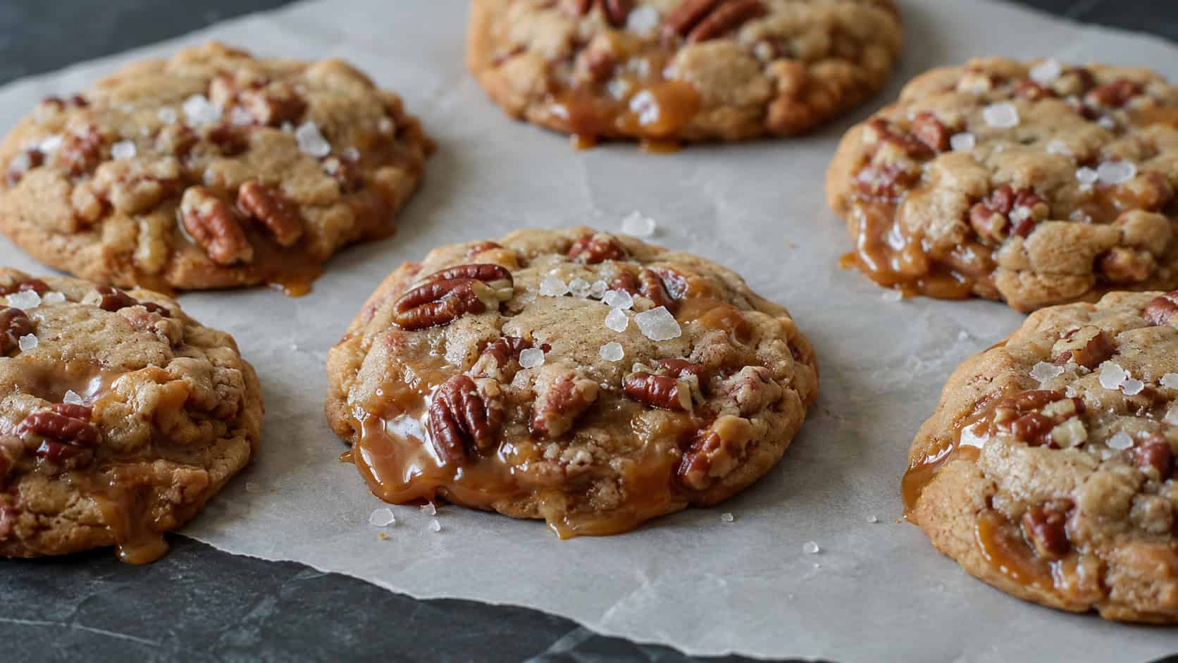 chewy salted caramel pecan cookies in a wide horizontal shot, oozing caramel and topped with flaky salt, arranged on parchment paper across a dark slate surface, soft natural light, shallow depth of field, landscape orientation