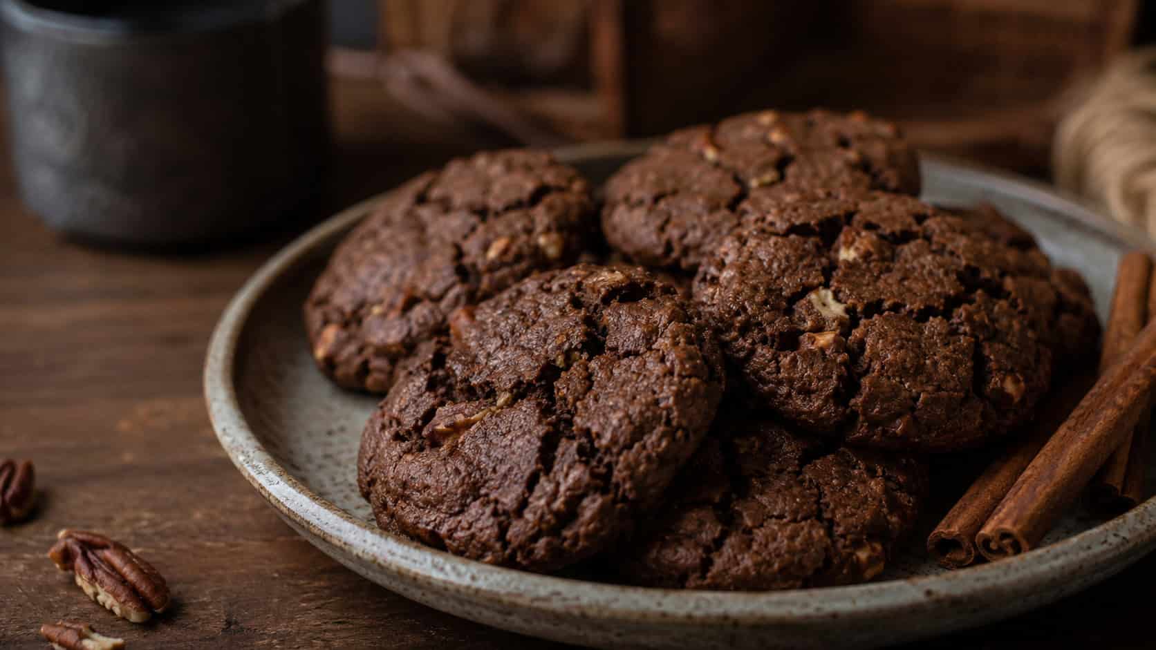 dark chewy molasses pecan cookies in a wide horizontal shot, crackled on top and arranged on a rustic ceramic plate with cinnamon sticks alongside, moody natural light, shallow depth of field, landscape orientation