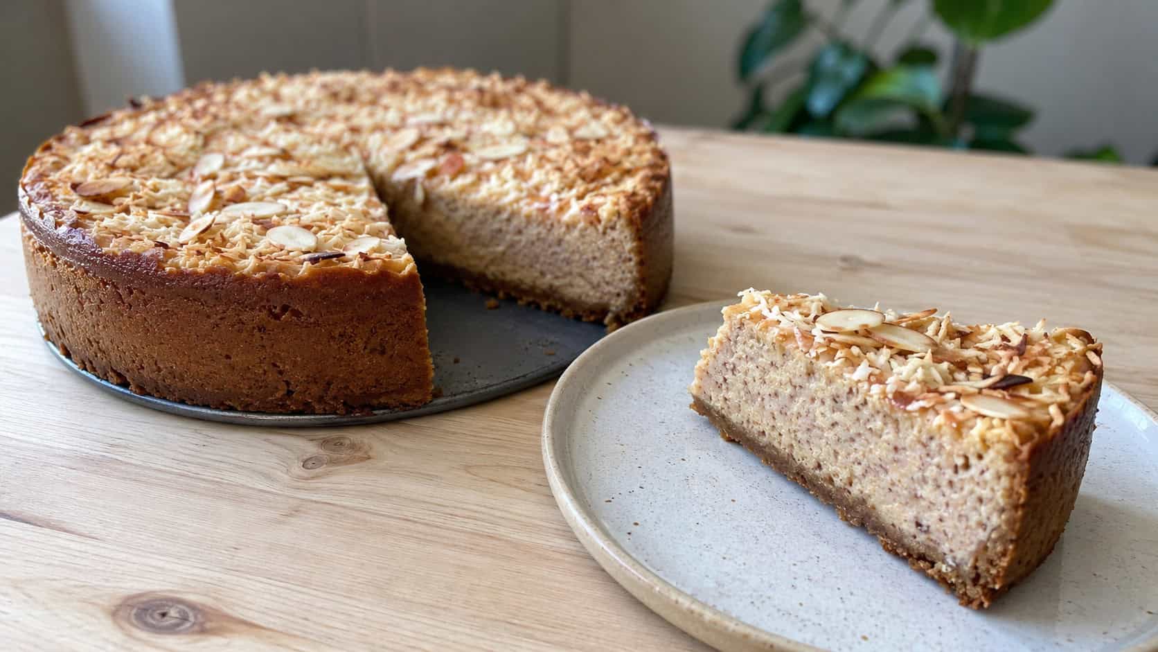 a cinnamon cheesecake topped with golden toasted coconut and almond slivers, one slice on a plate, in a wide horizontal shot on a light wooden table, natural light, shallow depth of field, landscape orientation