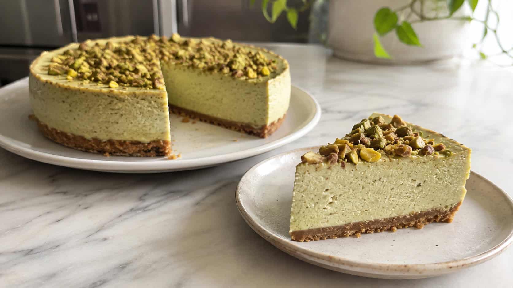 a faintly green cinnamon cheesecake topped with chopped pistachios, one slice on a plate, in a wide horizontal shot on a light marble counter, natural light, shallow depth of field, landscape orientation