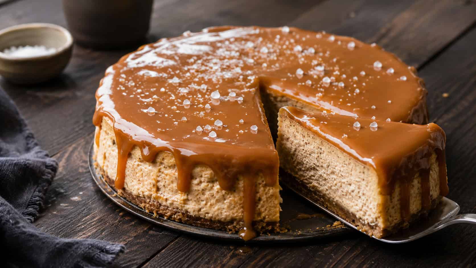 a cinnamon cheesecake covered in glossy salted caramel with flecks of sea salt on top, one slice pulled out, in a wide horizontal shot on a dark wooden table, natural light, shallow depth of field, landscape orientation