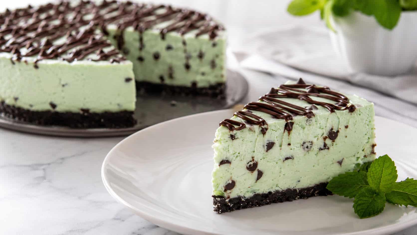 a pale mint-green cheesecake speckled with dark chocolate chips and a chocolate drizzle on top, one slice plated, on a cool white plate in a wide horizontal shot, on a clean table with a sprig of fresh mint, bright natural light, shallow depth of field, landscape orientation