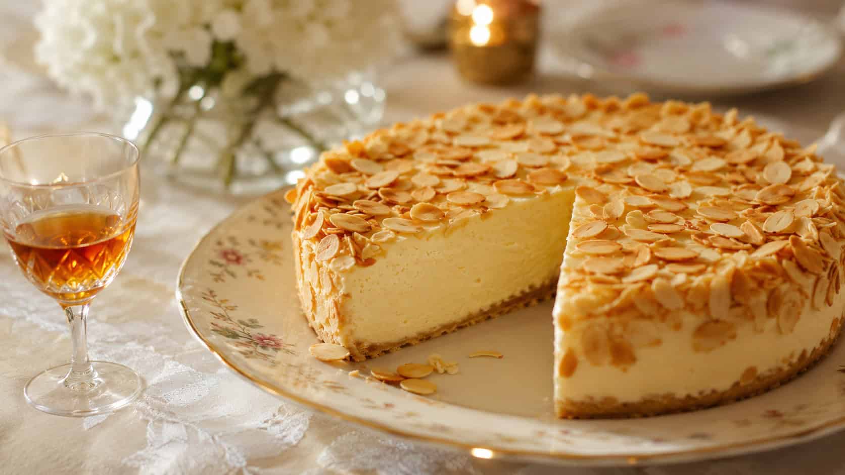 a smooth ivory almond cheesecake topped and edged with toasted sliced almonds, one slice cut, on a delicate china plate in a wide horizontal shot, on a refined table with a small glass of amber liqueur, soft elegant light, shallow depth of field, landscape orientation