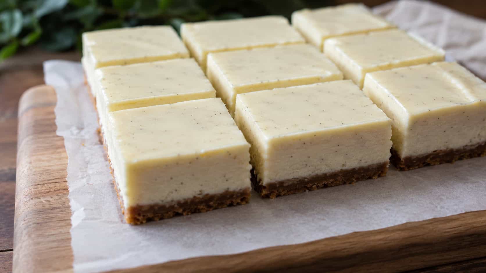 creamy vanilla bean cheesecake bars cut into neat squares on parchment, tiny black vanilla flecks visible, on a wooden board, wide horizontal shot, natural light, shallow depth of field, landscape orientation