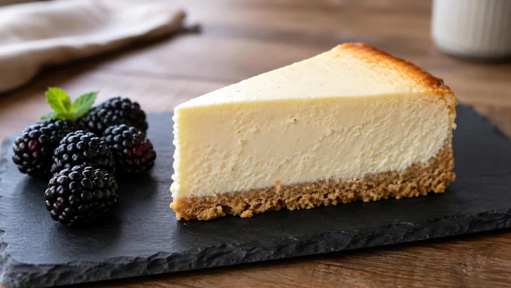 a low carb vanilla cheesecake slice with a golden almond crust on a slate plate, fresh blackberries beside it, on a wooden table, wide horizontal shot, natural light, shallow depth of field, landscape orientation