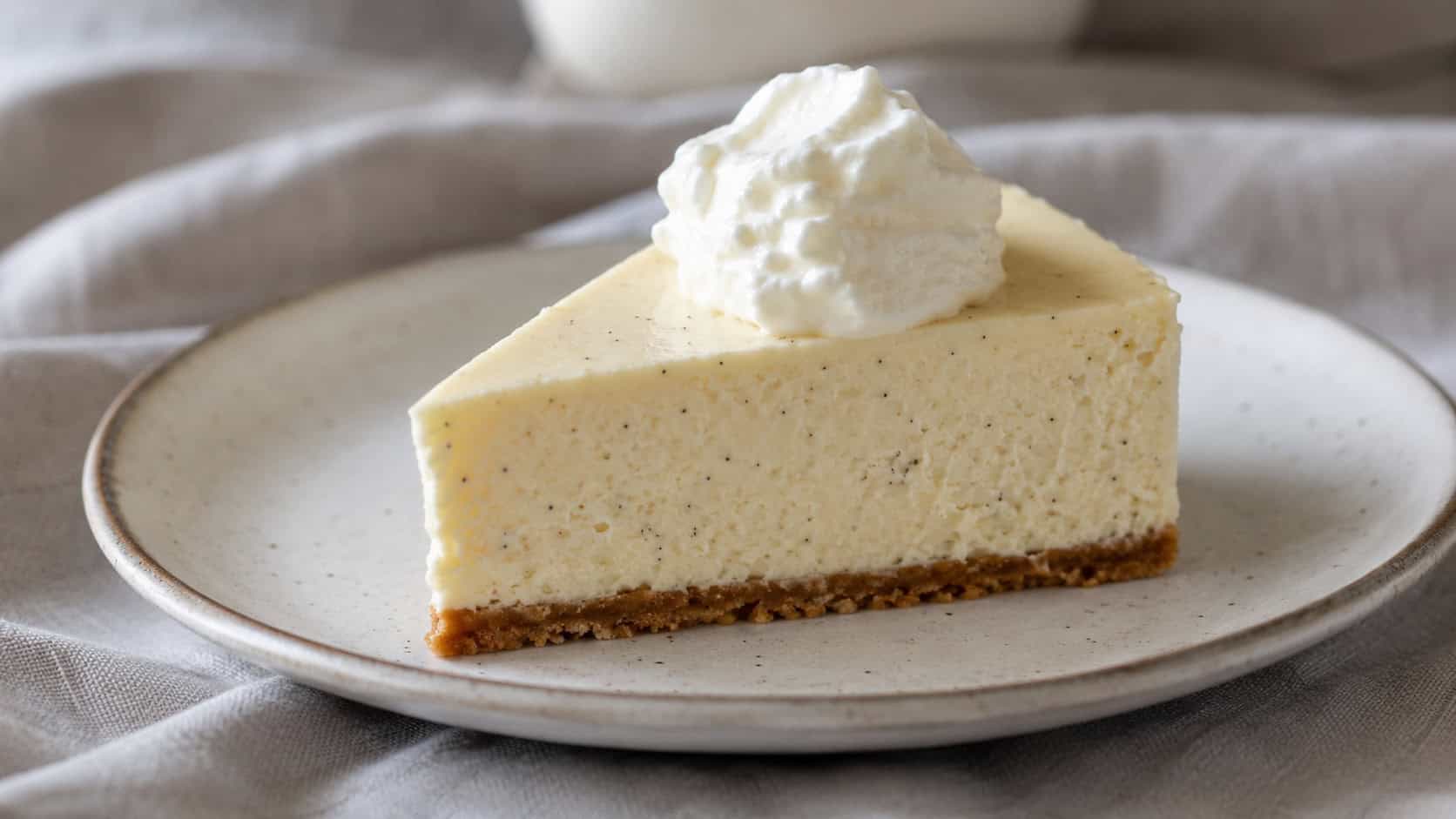 a creamy vanilla bean cheesecake slice flecked with tiny black vanilla seeds on a ceramic plate, dollop of whipped cream on top, on a soft linen cloth, wide horizontal shot, natural light, shallow depth of field, landscape orientation