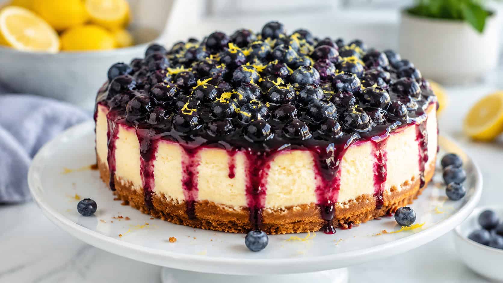 a lemon cheesecake topped with glossy blueberry compote dripping down the sides, fresh blueberries and lemon zest scattered around, on a white cake stand, wide horizontal shot, bright natural light, shallow depth of field, landscape orientation