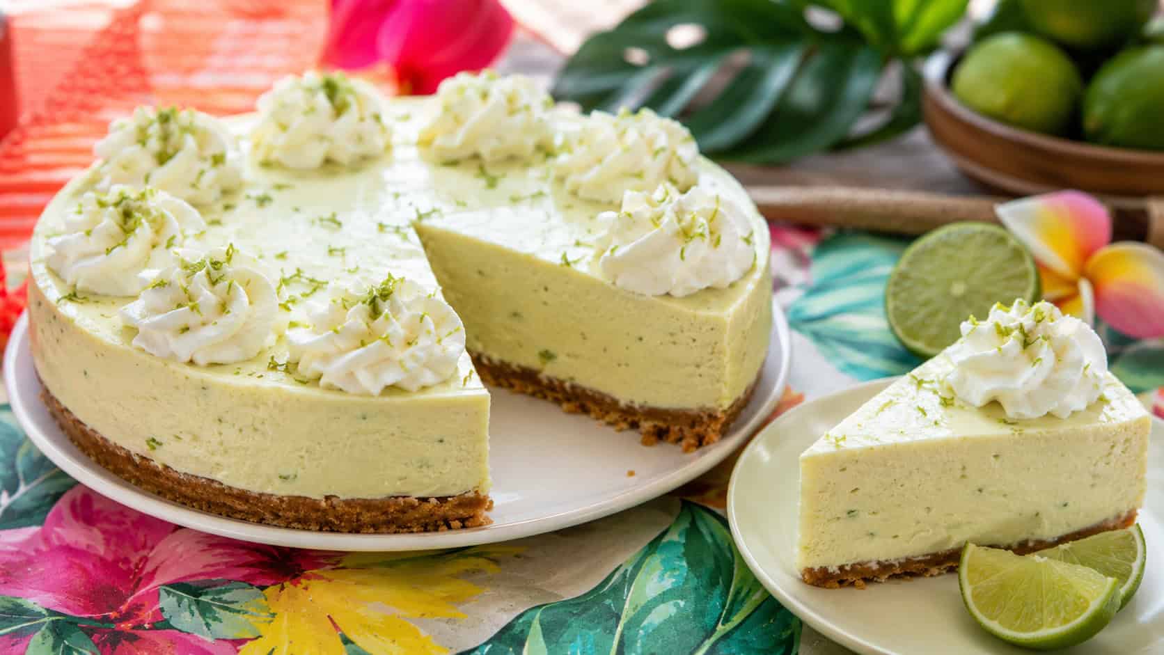 a pale green key lime cheesecake topped with whipped cream swirls and lime zest, a slice cut beside lime wedges, on a bright tropical-themed table, wide horizontal shot, bright natural light, shallow depth of field, landscape orientation