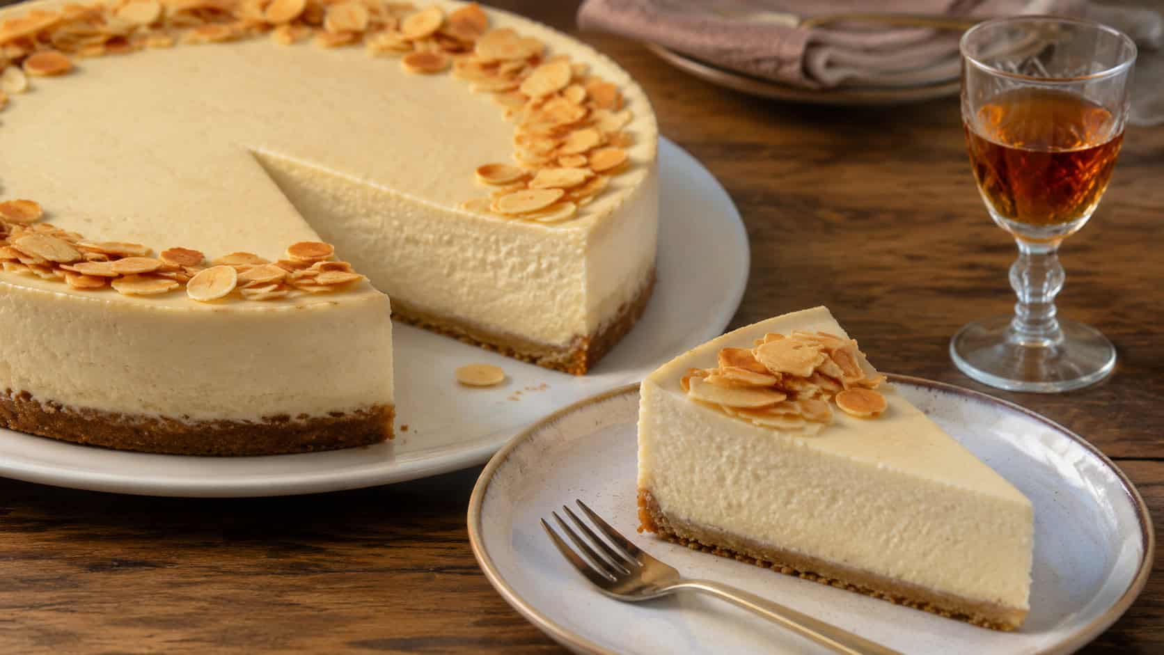 an amaretto cheesecake topped with toasted sliced almonds around the edges, a slice on a plate beside a small glass of liqueur, on a warm wooden table, wide horizontal composition, soft natural light, shallow depth of field, landscape orientation
