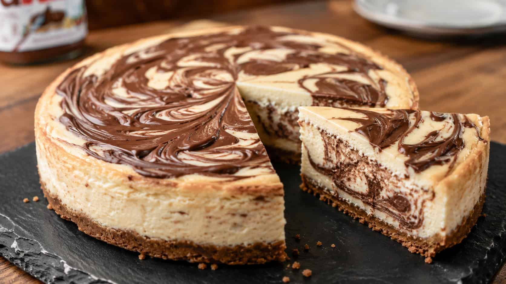 a Nutella swirl cheesecake with marbled chocolate ribbons across the top, one slice cut to reveal the swirl inside, on a dark slate board, wide horizontal shot, warm natural light, shallow depth of field, landscape orientation