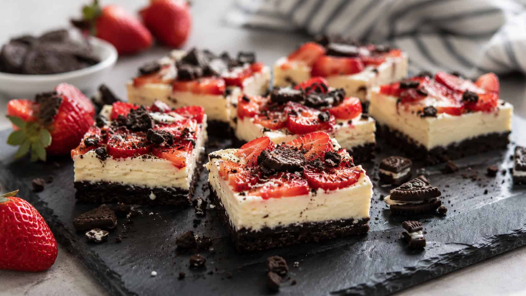 cheesecake bars on a dark chocolate cookie crust topped with fresh strawberry slices and cookie crumbs on a black slate board in a wide horizontal shot, whole berries and cookie pieces scattered around, natural light, shallow depth of field, landscape orientation