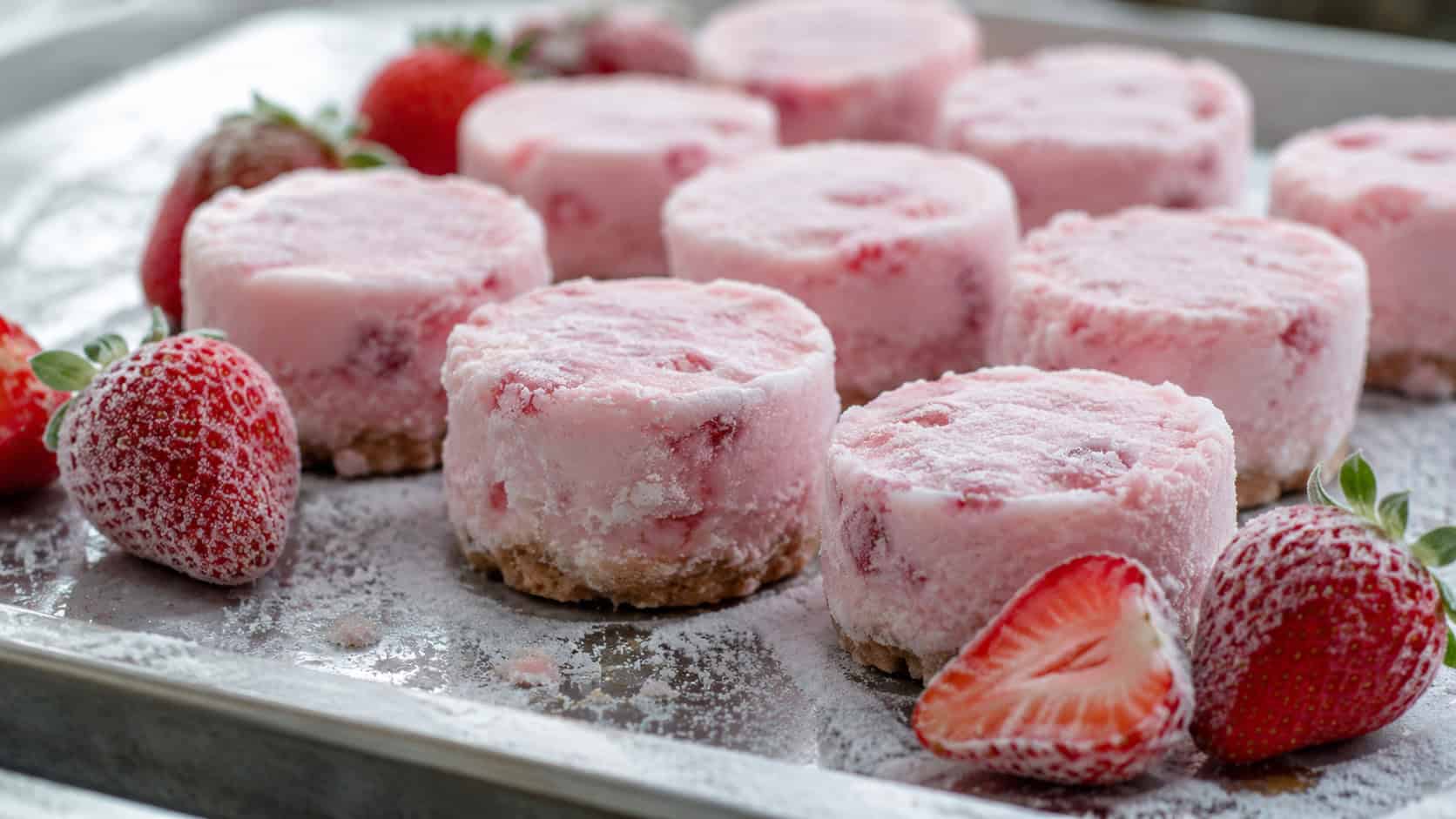 frosty pink frozen strawberry cheesecake bites on a chilled metal tray in a wide horizontal shot, fresh strawberries and a light dusting of frost around them, cool natural light, shallow depth of field, landscape orientation