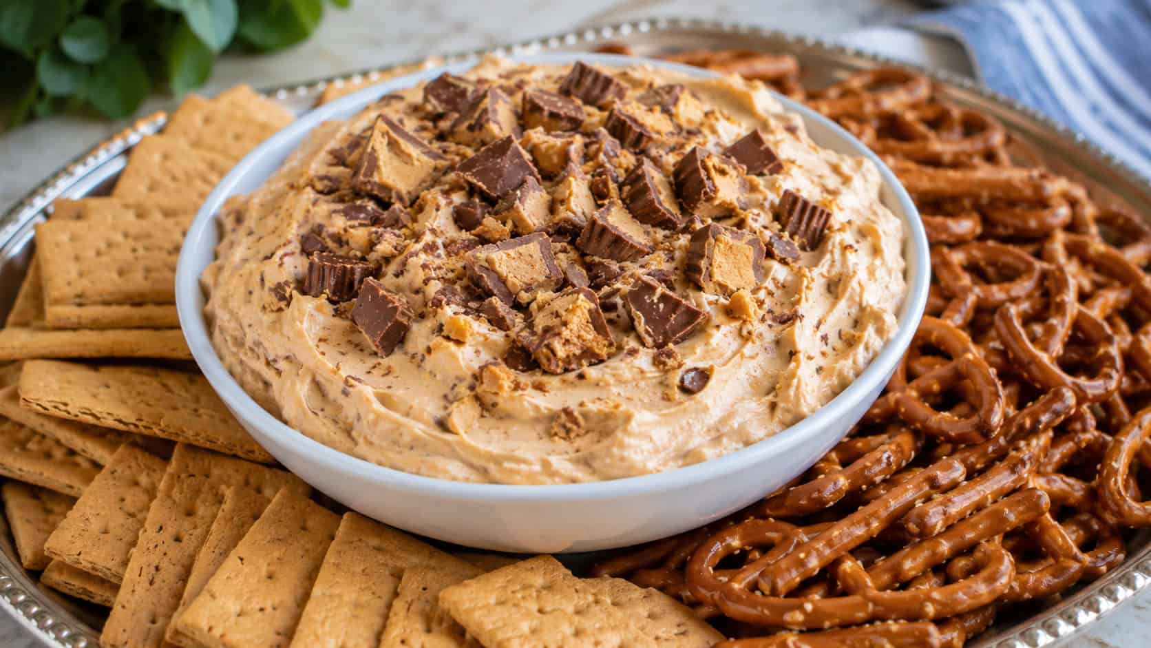 a bowl of creamy peanut butter cheesecake dip topped with chopped candy in a wide horizontal shot, surrounded by graham crackers and pretzels on a platter, natural light, shallow depth of field, landscape orientation
