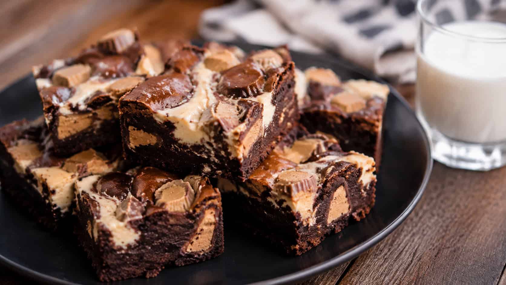 fudgy cheesecake-swirled brownies studded with peanut butter candies in a wide horizontal shot, stacked on a dark plate with a glass of milk, natural light, shallow depth of field, landscape orientation