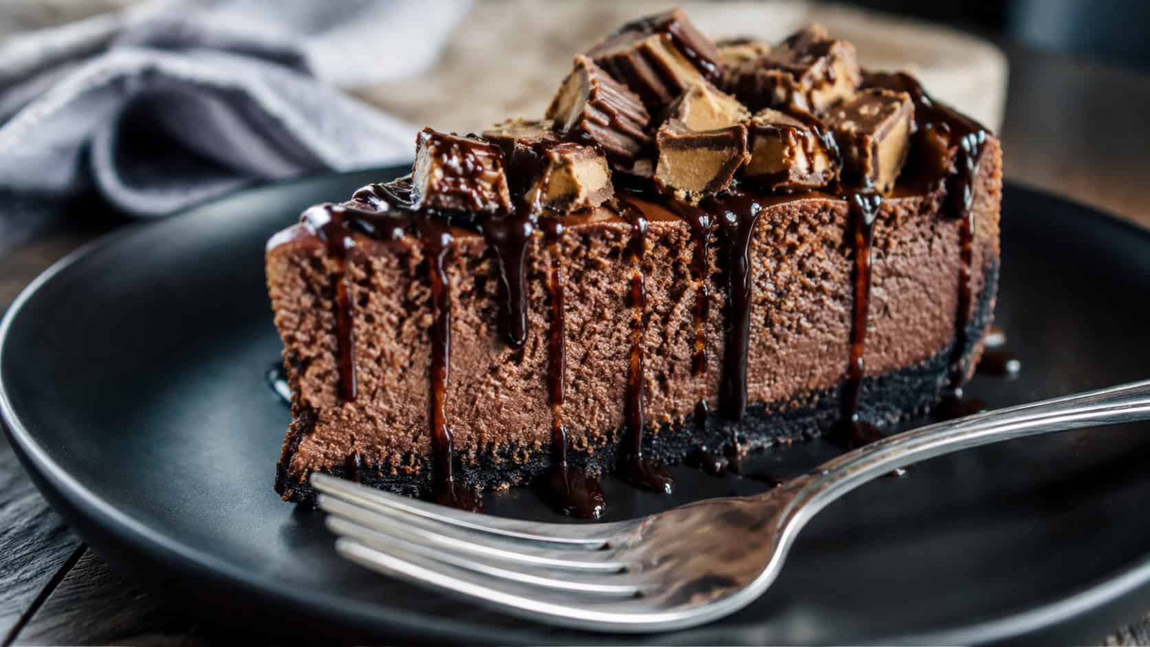 a dark chocolate cheesecake with ganache drizzle and chopped peanut butter cups in a wide horizontal shot, on a black plate with a fork, natural light, shallow depth of field, landscape orientation