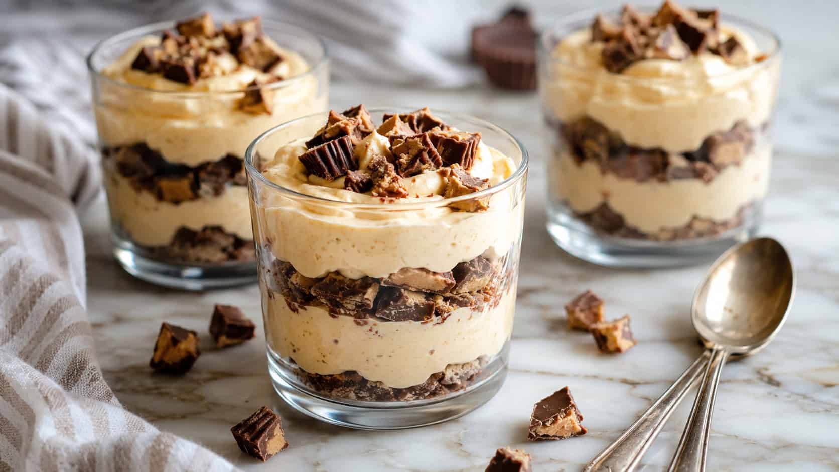 glasses of fluffy peanut butter cheesecake mousse layered with crushed peanut butter cups in a wide horizontal shot, on a marble surface with spoons, natural light, shallow depth of field, landscape orientation