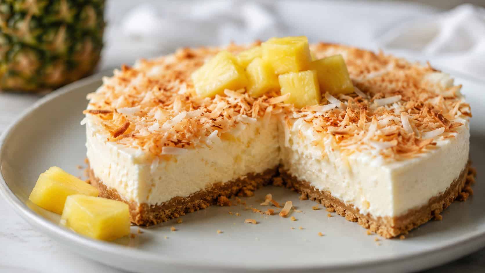 a no-bake pineapple coconut cheesecake topped with toasted coconut in a wide horizontal shot, on a pale plate with pineapple chunks, soft natural light, shallow depth of field, landscape orientation