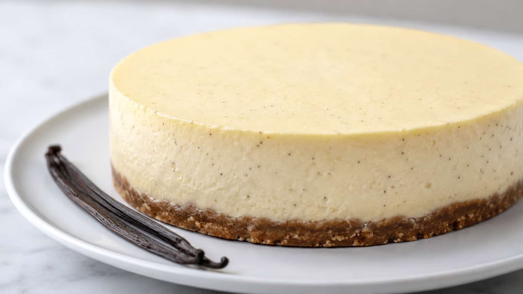a pale creamy vanilla bean cheesecake flecked with tiny black vanilla specks in a wide horizontal shot, a spent vanilla pod beside it, soft white plate, bright clean natural light, shallow depth of field, landscape orientation