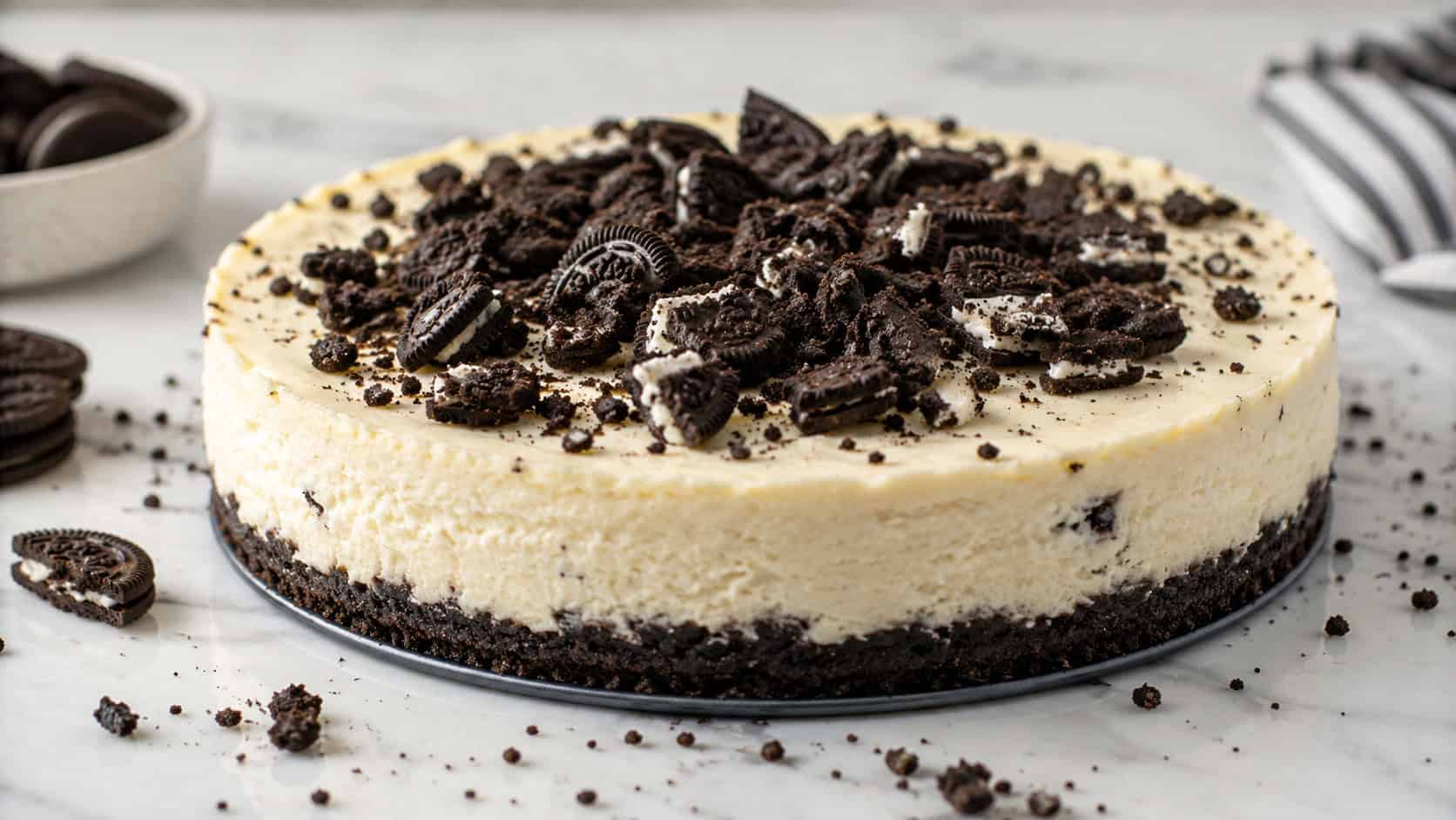 a creamy cheesecake on a dark cookie crumb crust with chocolate sandwich cookie pieces on top in a wide horizontal shot, crumbs scattered around, light marble counter, soft natural light, shallow depth of field, landscape orientation