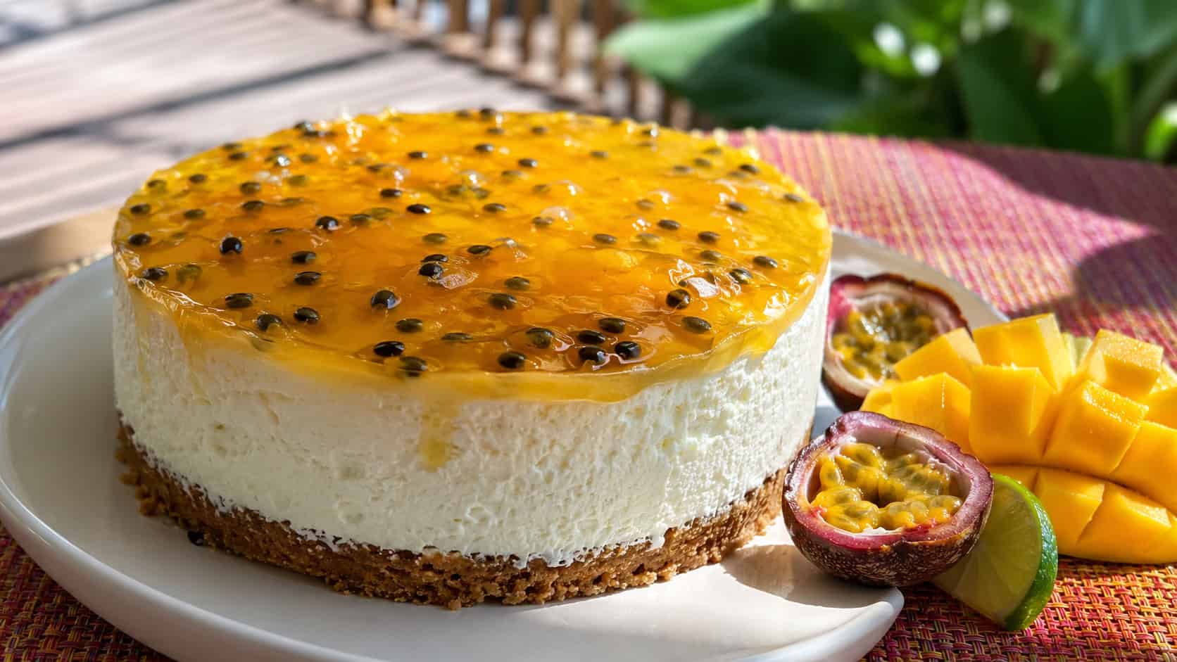 a creamy cheesecake topped with golden mango glaze and passionfruit seeds in a wide horizontal shot, tropical fruit slices beside it, bright woven placemat, sunny natural light, shallow depth of field, landscape orientation