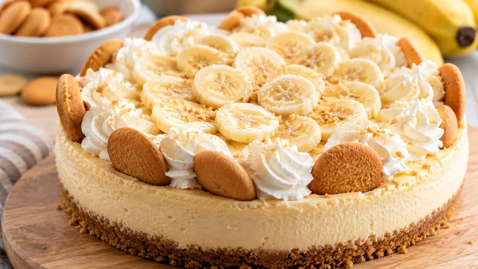 a creamy cheesecake topped with swirls of whipped cream and fresh banana slices in a wide horizontal shot, vanilla wafers tucked around the edge, light wooden board, soft natural light, shallow depth of field, landscape orientation