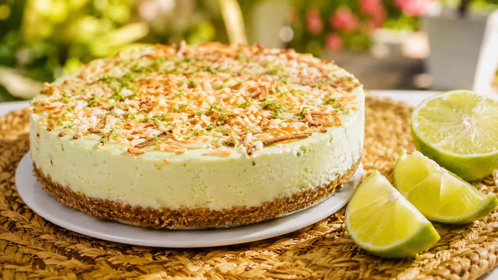 a pale green-tinted lime cheesecake topped with toasted golden coconut and lime zest in a wide horizontal shot, lime wedges on the side, bright woven mat, sunny natural light, shallow depth of field, landscape orientation