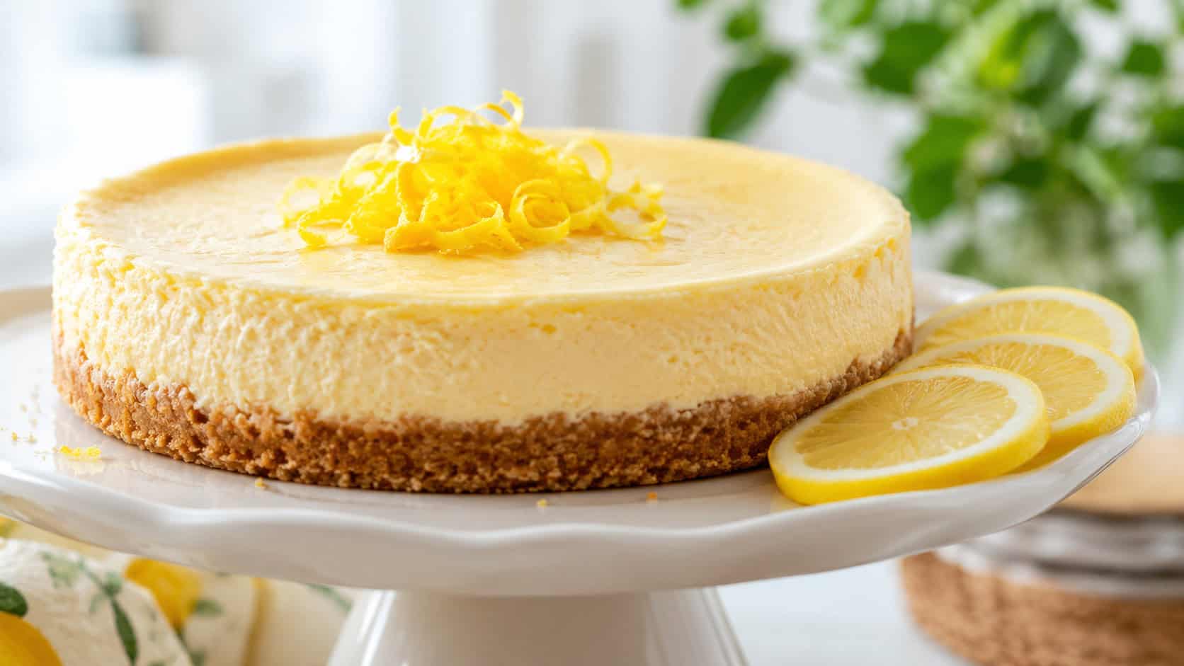 a pale golden lemon cheesecake garnished with curls of lemon zest in a wide horizontal shot, thin lemon slices on the side, white ceramic stand, fresh bright daylight, shallow depth of field, landscape orientation