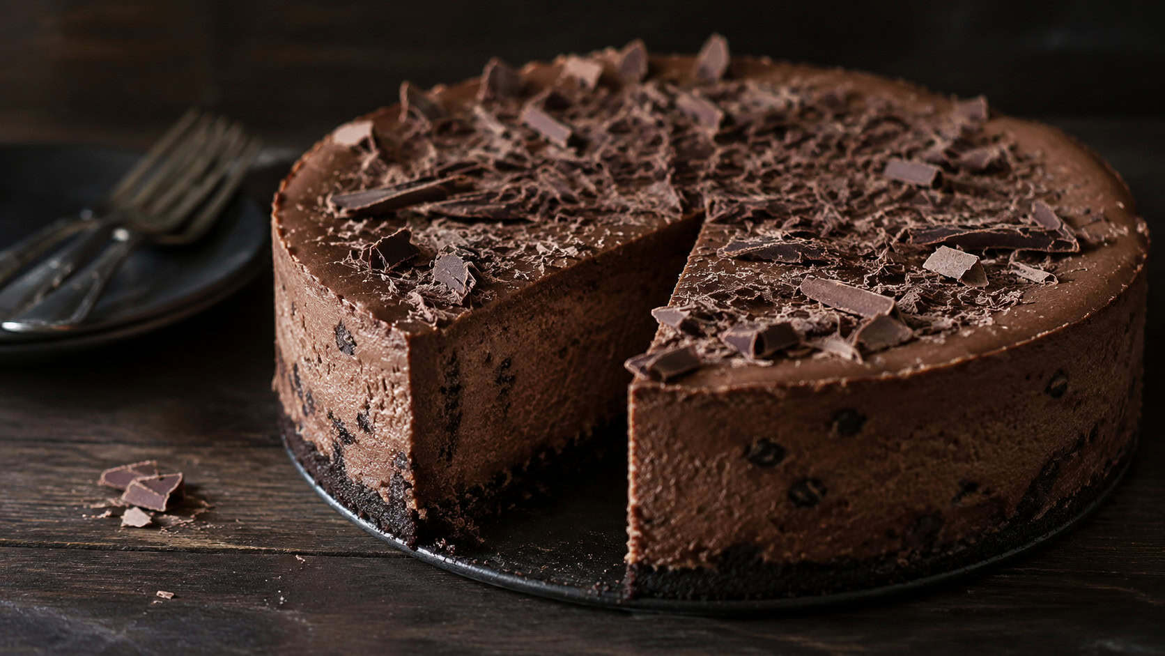 a rich dark chocolate cheesecake speckled with chocolate chips in a wide horizontal shot, chocolate shavings on top, dark wooden surface, moody soft natural light, shallow depth of field, landscape orientation