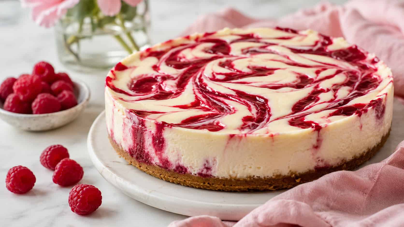 a creamy cheesecake with bright red raspberry ripples swirled across the top in a wide horizontal shot, fresh raspberries scattered nearby, soft pink linen, gentle natural light, shallow depth of field, landscape orientation