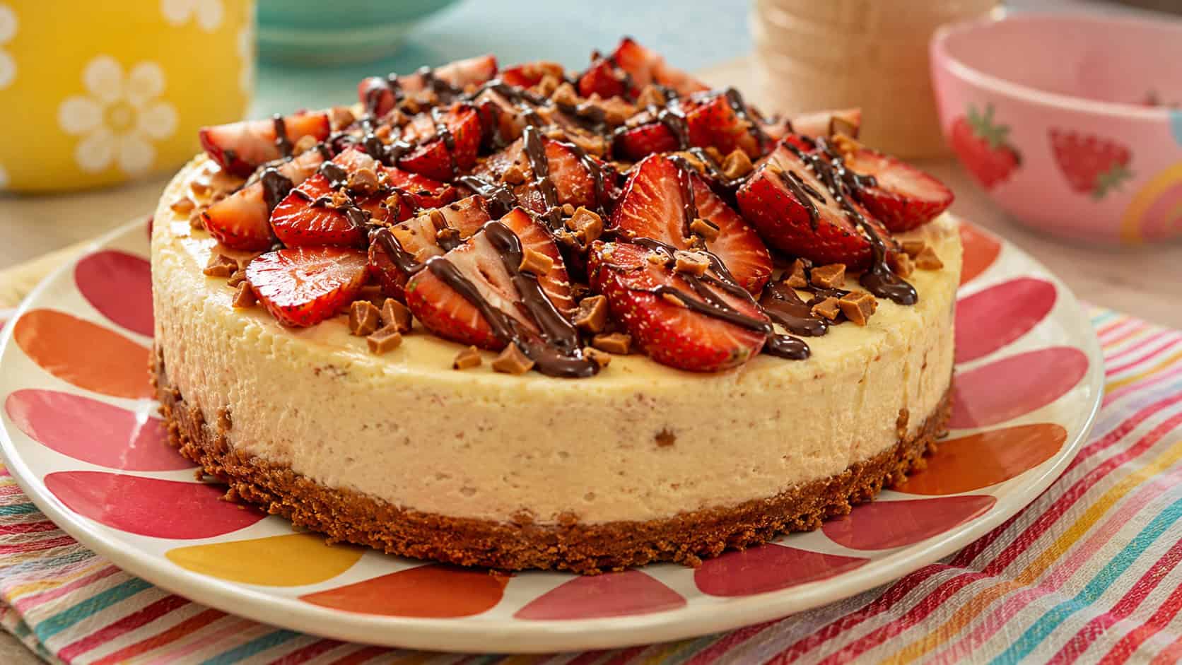 a banana cheesecake topped with fresh strawberries, chocolate drizzle, and toffee in a wide horizontal shot, on a cheerful retro plate with a striped napkin, bright playful light, shallow depth of field, landscape orientation