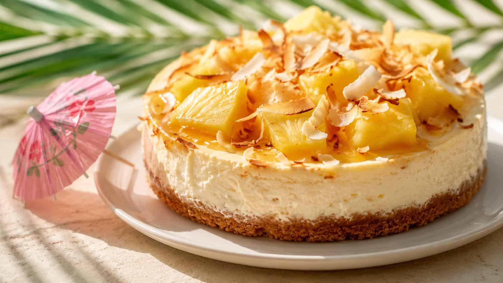 a tropical cheesecake topped with toasted coconut and pineapple in a wide horizontal shot, a paper umbrella on the side, set on a bright table with palm shadows, sunny natural light, shallow depth of field, landscape orientation
