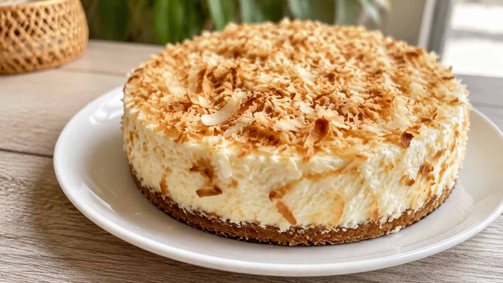 a coconut cheesecake heaped with toasted coconut flakes in a wide horizontal shot, set on a white plate on a light wood table, bright natural light, shallow depth of field, landscape orientation