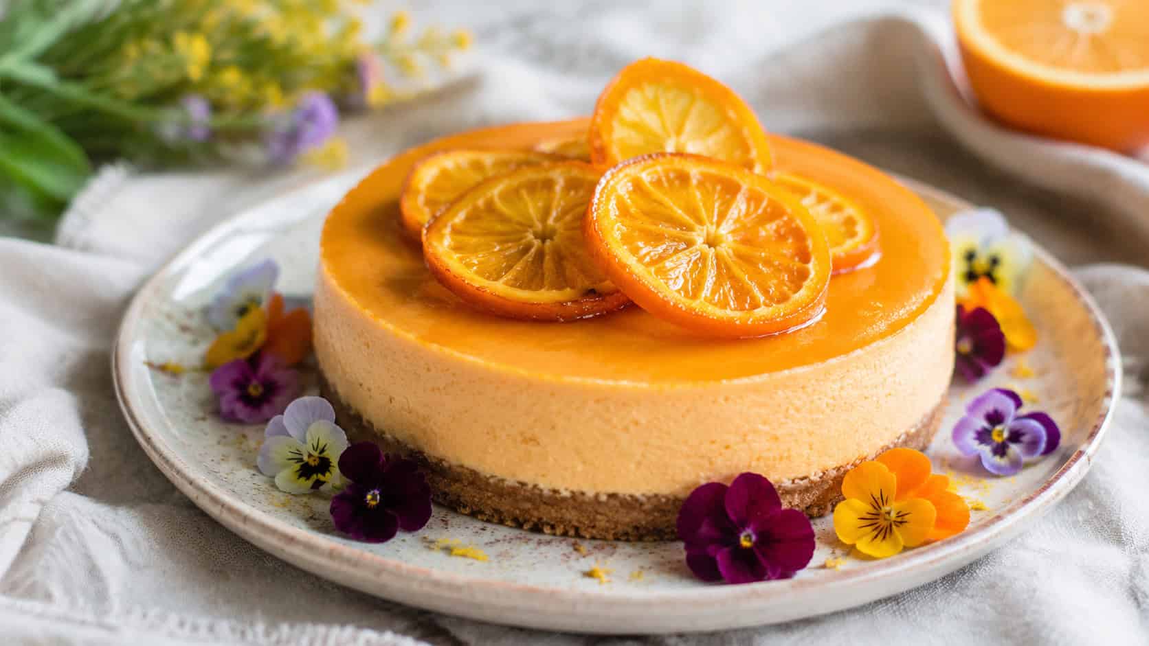 a delicate orange cheesecake topped with candied orange slices in a wide horizontal shot, edible flowers around the plate, set on a pale linen surface, soft natural light, shallow depth of field, landscape orientation