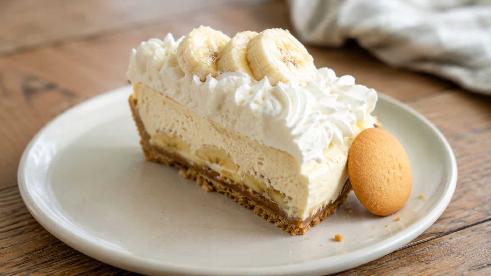 a banana cheesecake topped with whipped cream and fresh banana slices in a wide horizontal shot, a vanilla wafer on the side, set on a cream plate on a wood table, natural light, shallow depth of field, landscape orientation