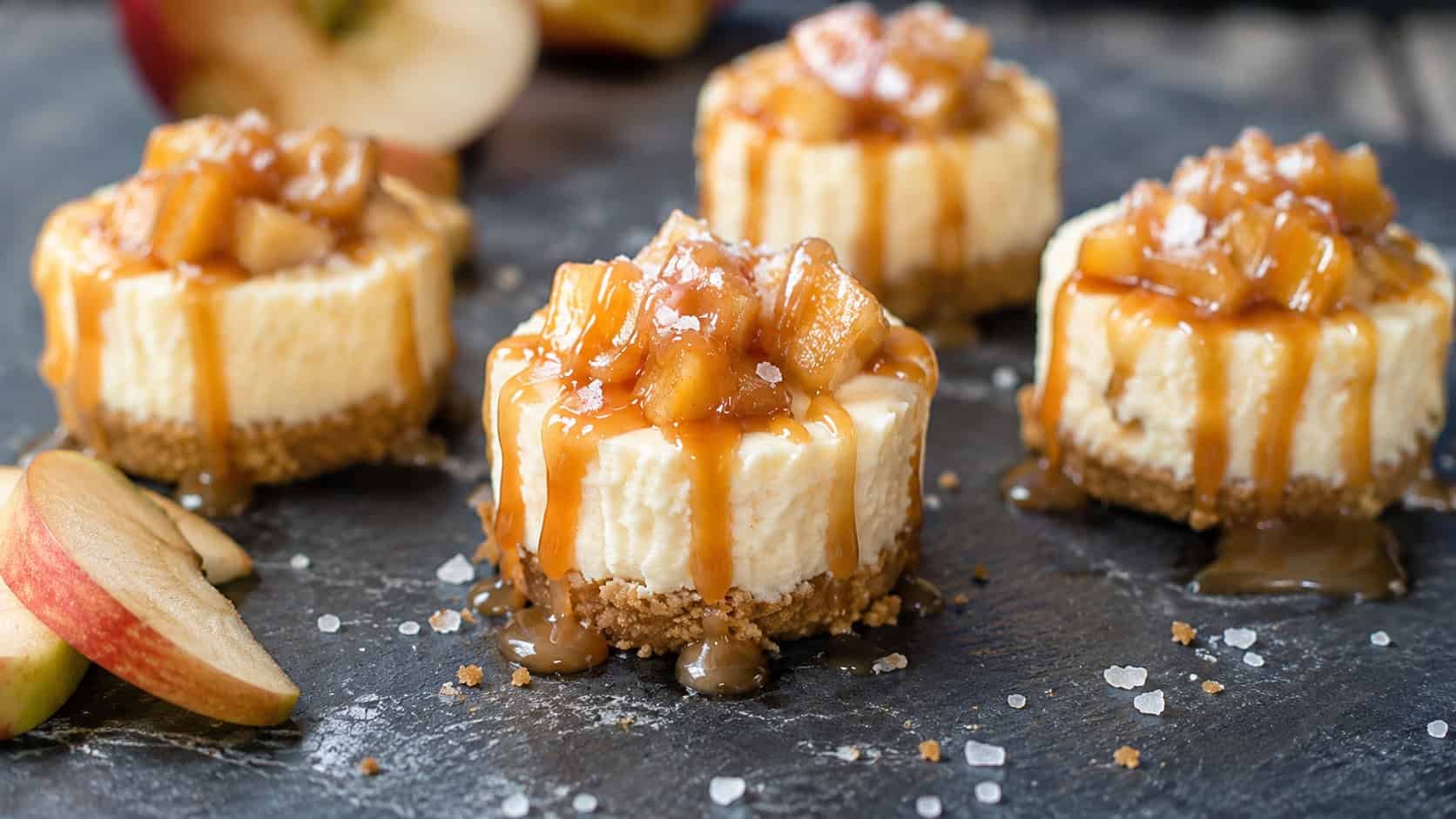 mini cheesecakes with caramel apples and flaky salt drizzled generously, in a wide horizontal shot on a dark slate board, natural light, shallow depth of field, landscape orientation