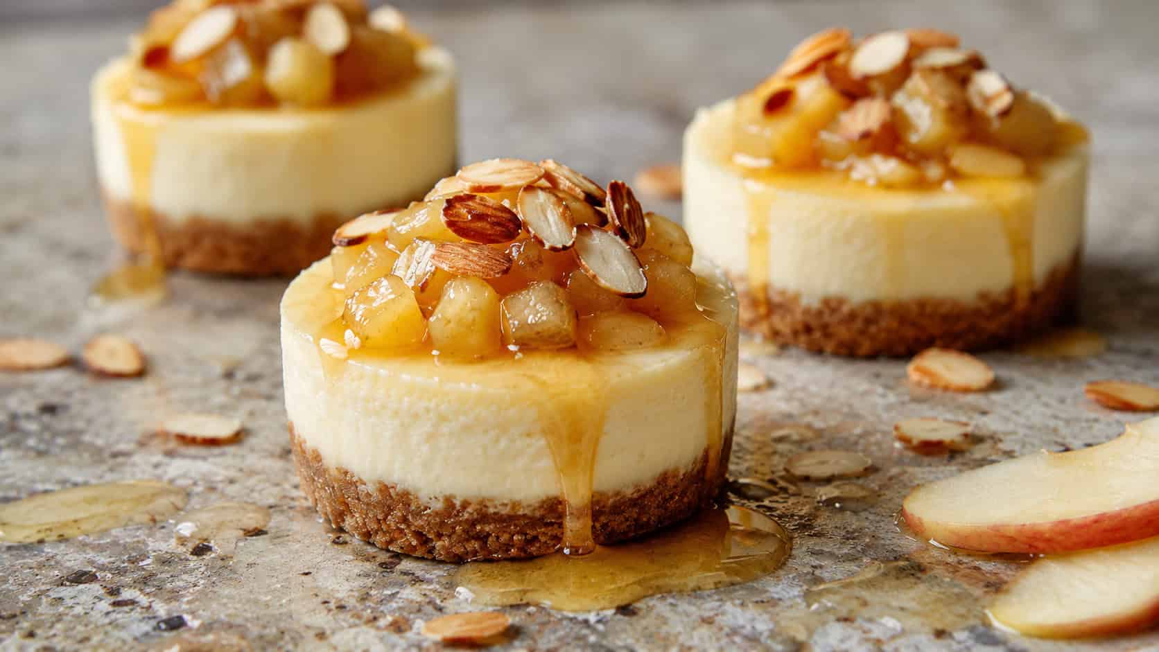 honey cheesecakes topped with apples and toasted sliced almonds, in a wide horizontal shot on a stone surface drizzled with honey, natural light, shallow depth of field, landscape orientation