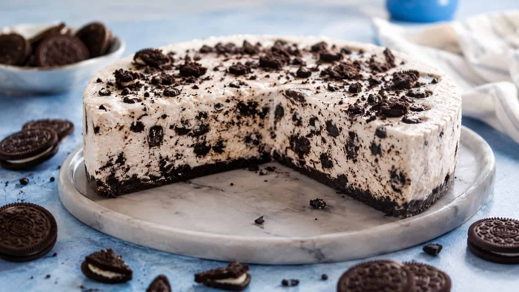 a frozen cookies and cream cheesecake on a wide chilled platter in a horizontal shot, a firm icy slice packed with dark cookie pieces and a frosty sheen, scattered cookies on a cool blue table, bright daylight, shallow depth of field, landscape orientation