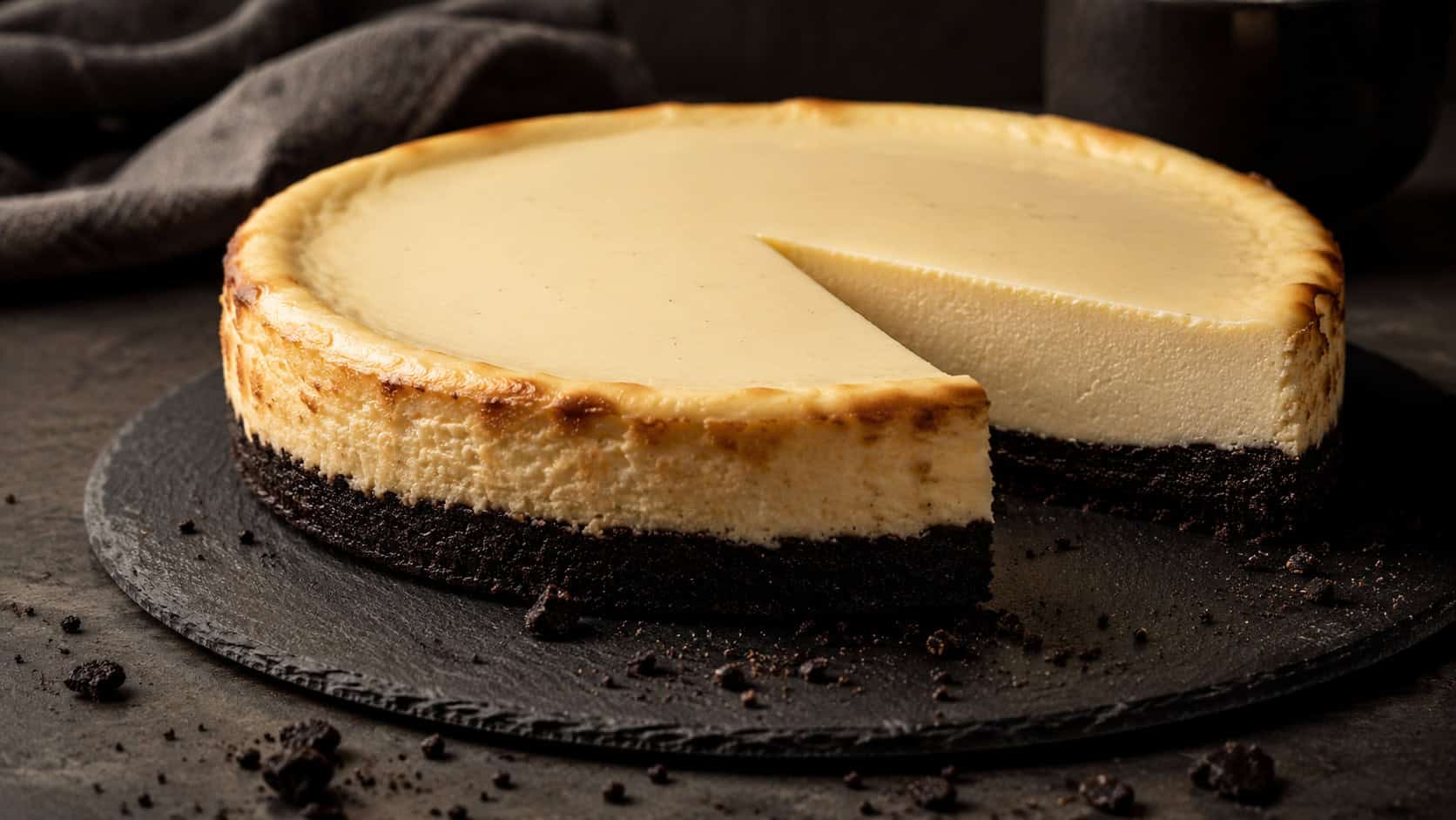 a vanilla cheesecake with a thick dark cookie crust on a slate board in a wide horizontal shot, one clean slice removed to reveal the deep crust layer, cookie crumbs scattered nearby on a moody dark counter, warm directional light, shallow depth of field, landscape orientation
