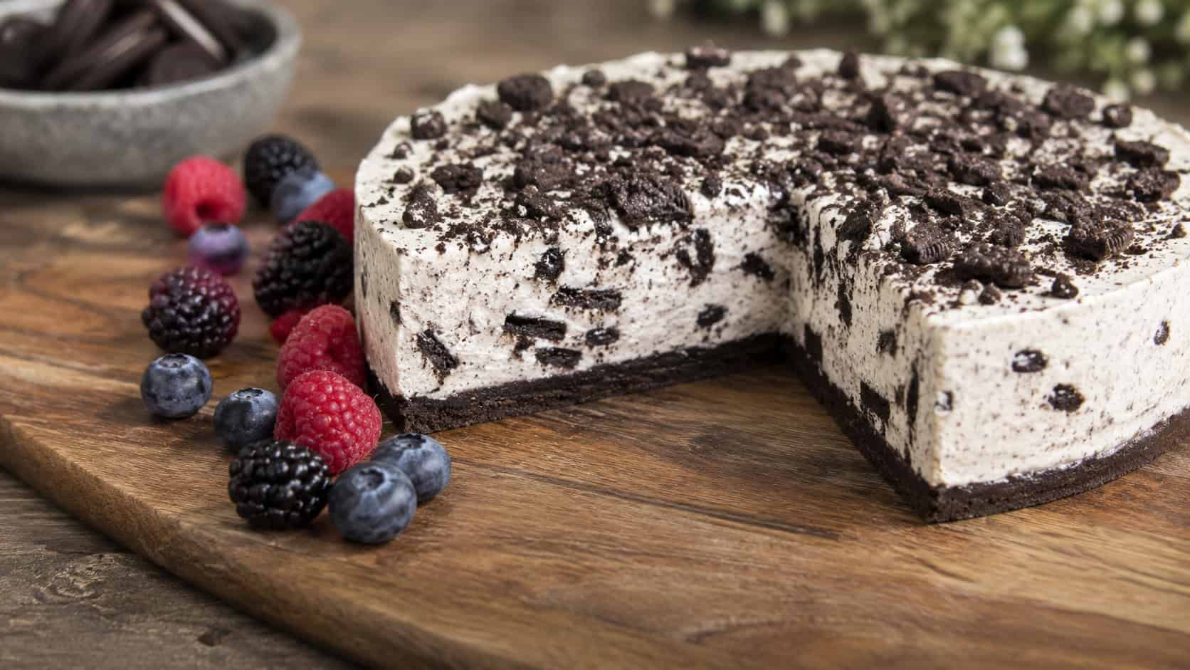 a dairy-free cookies and cream cheesecake on a wide wooden board in a horizontal shot, a smooth pale slice flecked with dark cookie pieces and crushed cookie topping, fresh berries scattered alongside on a rustic table, soft natural light, shallow depth of field, landscape orientation