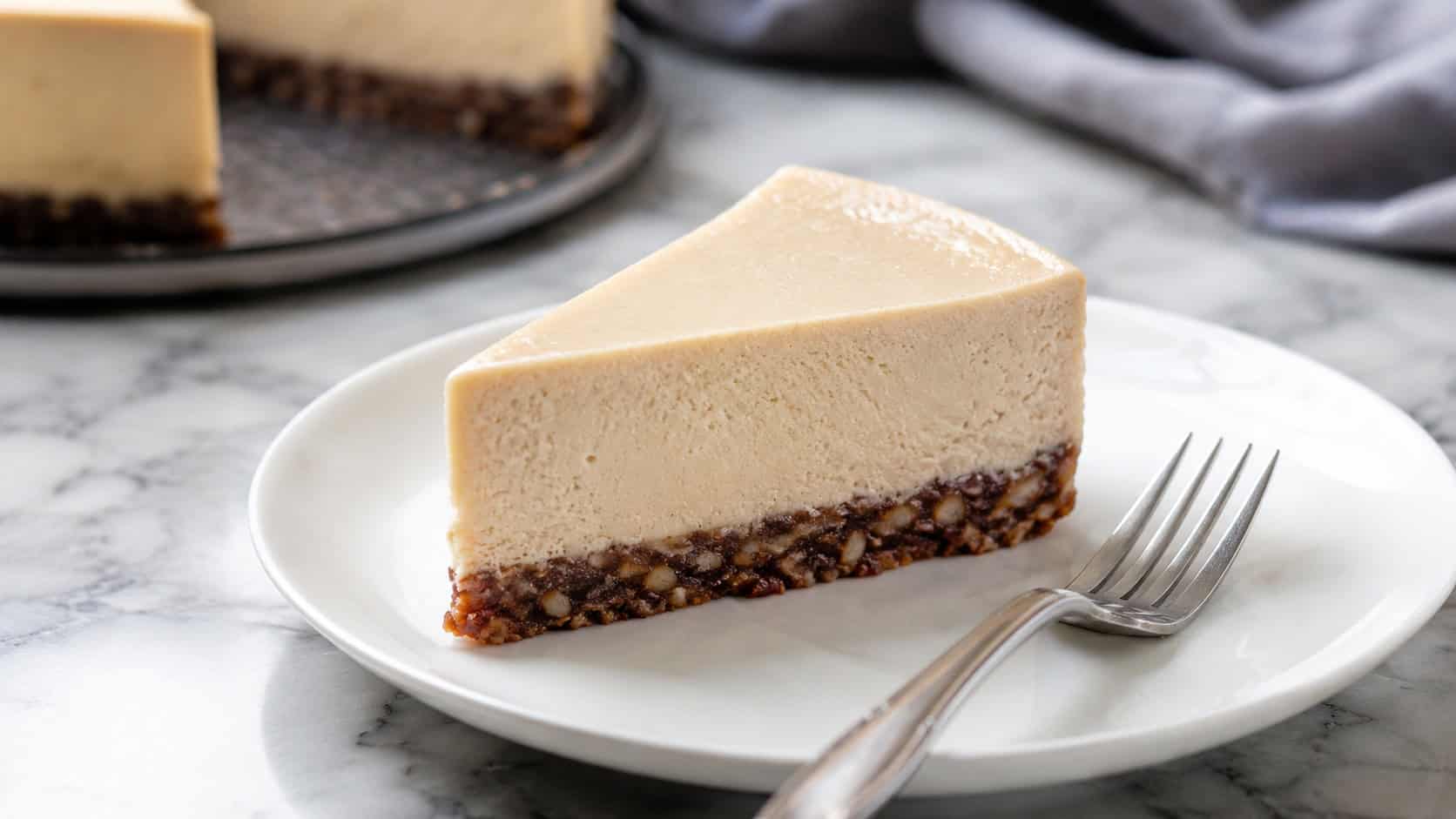 a classic cashew cheesecake in a wide horizontal shot, smooth pale slice on a date-nut crust, on a white plate with a fork, on a marble counter, natural light, shallow depth of field, landscape orientation