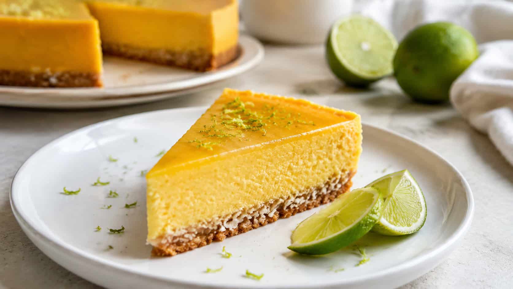 a mango lime cheesecake in a wide horizontal shot, bright golden slice topped with lime zest on a coconut crust, on a white plate with lime wedges, on a light counter, natural light, shallow depth of field, landscape orientation