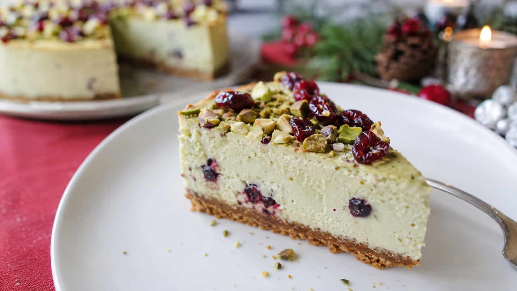 a pistachio cranberry cheesecake in a wide horizontal shot, pale green slice topped with red cranberries and chopped pistachios, on a white plate, on a festive table, natural light, shallow depth of field, landscape orientation
