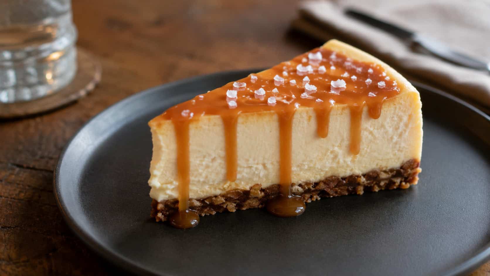 a salted caramel cheesecake in a wide horizontal shot, creamy slice with golden caramel drizzle and flaky salt on a nut crust, on a dark plate, on a wooden table, natural light, shallow depth of field, landscape orientation